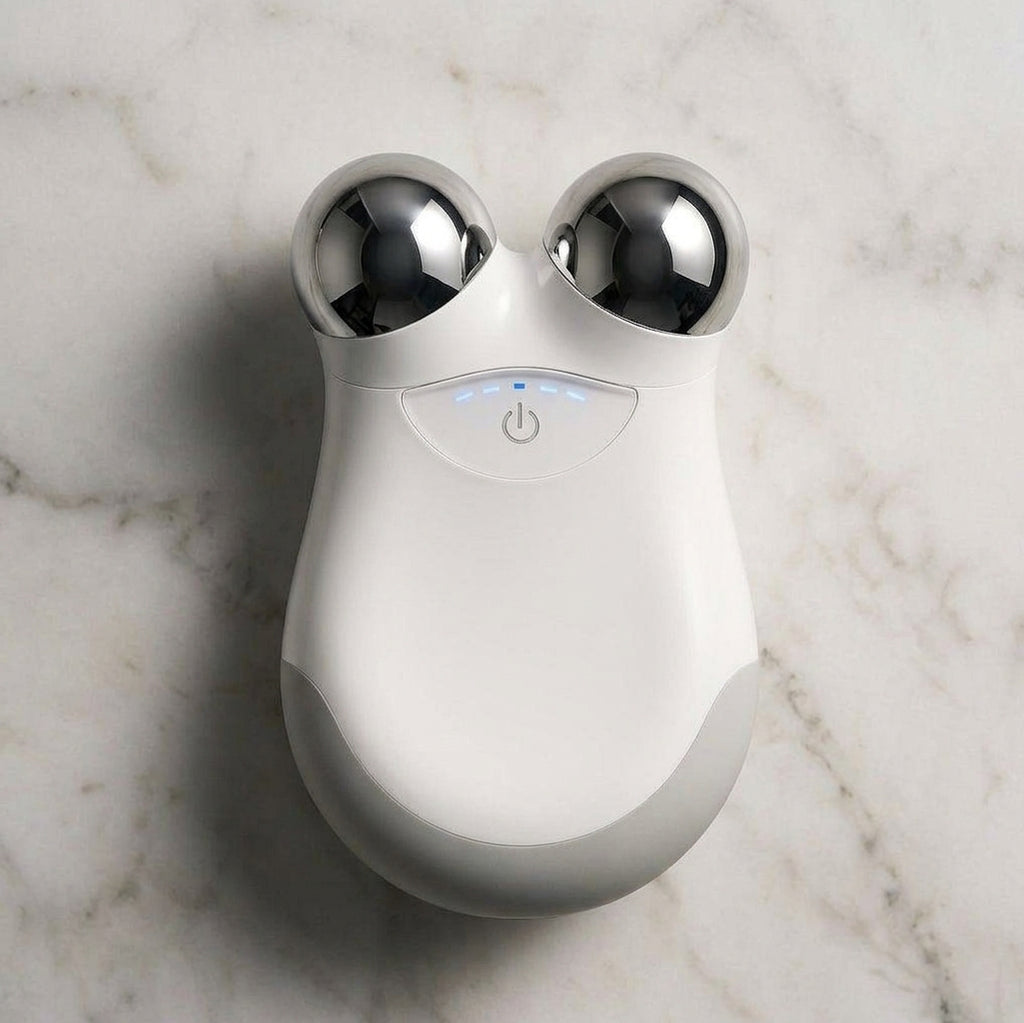 A white and gray handheld beauty device with two metallic silver spherical heads, resting on a white marble surface.
