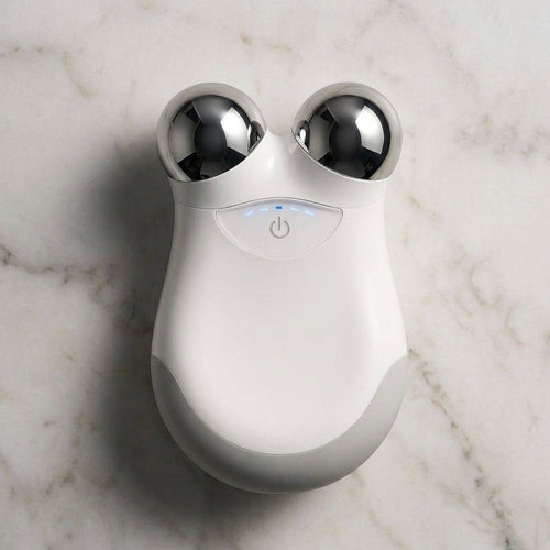 A white and gray handheld beauty device with two metallic silver spherical heads, resting on a white marble surface.
