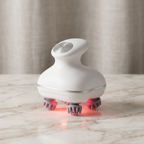 A white handheld scalp massage device with pink silicone massage bristles and a red light therapy base, resting on a light marble countertop with a soft beige curtain background.