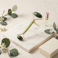 A jade roller tool with green stone ends and gold-tone metal accents rests on a white marble tray, styled with eucalyptus leaves, small white flowers, a small bottle of oil, and a cream-colored box on a light fabric background.